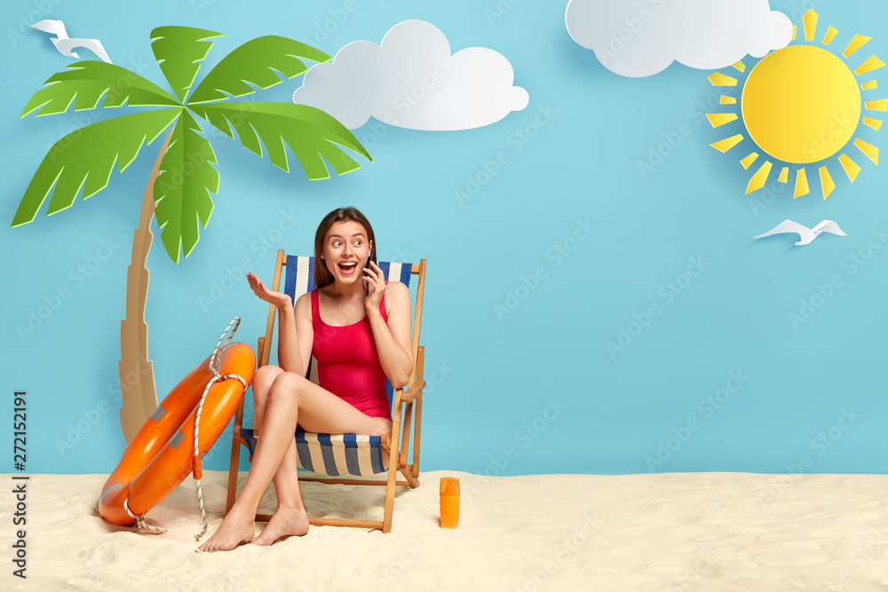 Happy excited woman in red swimwear, relaxes on deckchair at sandy beach sea coast, talks on cellphone, shares impressions with friend about nice rest, enjoys sunny day, uses sun protection cream