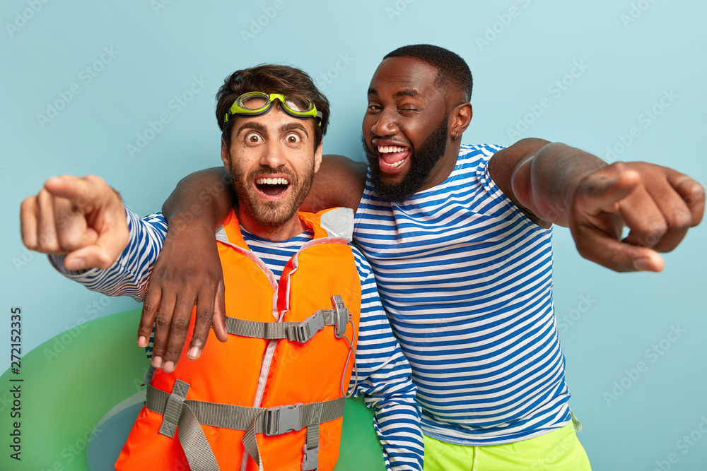 Happy diverse guys embrace and point straightly at camera, have fun at beach, pose with life preserver, lifejacket, being in high spirit during sunny hot day, notice extreme parachute jumper - obrazy, fototapety, plakaty 