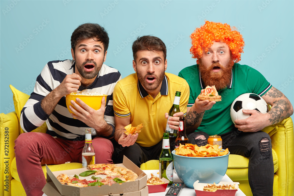 Three astonished male friends stare at camera, eat tasty pizza, popcorn, crisps, drink cold beer, watch football championship, being very emotional, sit on sofa at home against blue background Three astonished male friends stare at camera, eat tasty pizza, popcorn, crisps, drink cold beer, watch football championship, being very emotional, sit on sofa at home against blue background