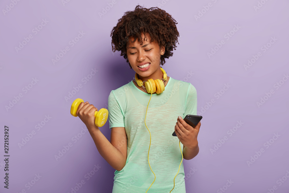 Sporty woman lifts weight, engaged in fitness, does exercises indoor, listens music with earphones and smartphone during training, wears t shirt. Dark skinned female has aerobics or workout in gym Sporty woman lifts weight, engaged in fitness, does exercises indoor, listens music with earphones and smartphone during training, wears t shirt. Dark skinned female has aerobics or workout in gym