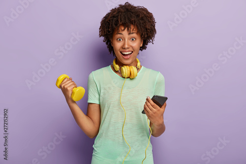 Delighted ethnic woman chats on mobile phone, listens music during jogigng exercises, warms up, holds dumbbell, uses headphones and mobile phone, models against purple background, attends health club