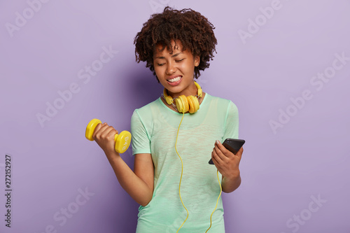 Sporty woman lifts weight, engaged in fitness, does exercises indoor, listens music with earphones and smartphone during training, wears t shirt. Dark skinned female has aerobics or workout in gym