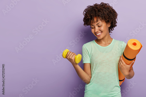 Healthy fitness girl lifts weight dumbbell, works to have biceps, carries rolled karemat, warms up before workout, has happy expression, isolated on purple wall. Wellness and recreation concept photo
