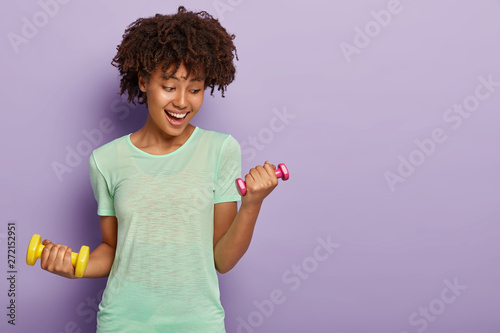 Sporty glad girl does biceps exercises with two small dumbbells, wants to have muscular arms, dressed casually, enjoys workout, isolated on purple wall. Energetic fitness instructor. Healthy lifestyle