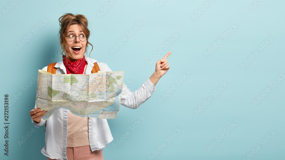 © wayhome.studio - Positive female traveler holds paper map, shows direction, being lost in unknown place, wears round spectacles, red bandana, points at free space, has glad look, isolated over blue background © wayhome.studio - Positive female traveler holds paper map, shows direction, being lost in unknown place, wears round spectacles, red bandana, points at free space, has glad look, isolated over blue background