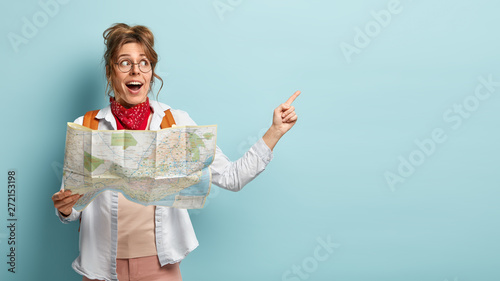 Positive female traveler holds paper map, shows direction, being lost in unknown place, wears round spectacles, red bandana, points at free space, has glad look, isolated over blue background photo