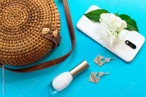 Straw stylish modern women's summer bag and phone, earrings and white jasmine flower on a blue background. Summer vacation concept