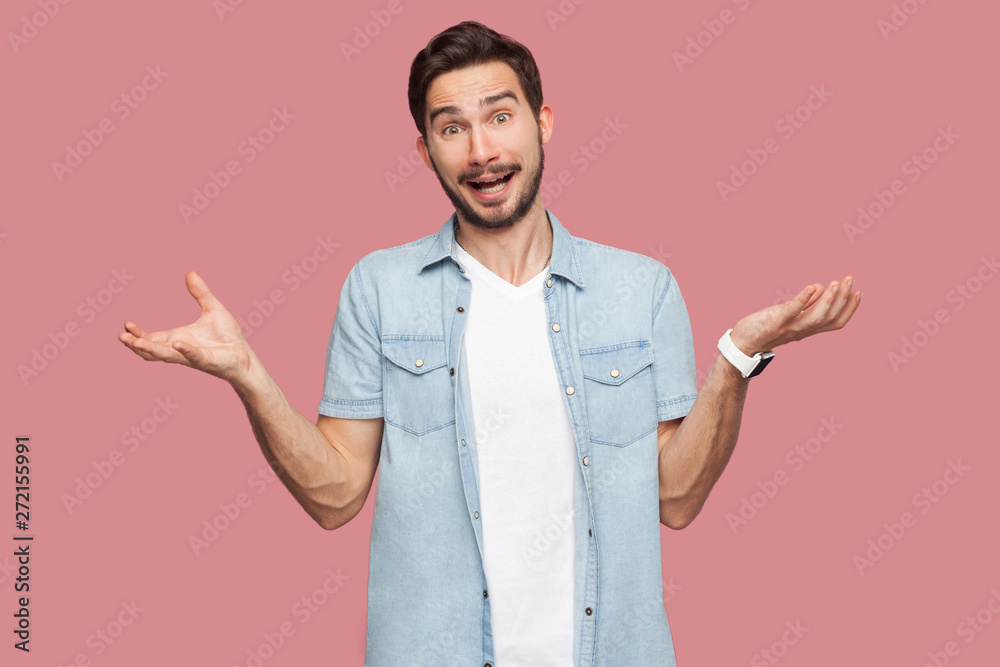 Portrait of surprised handsome bearded young man in blue casual style shirt standing with raised arms and looking at camera with amazed face. indoor studio shot, isolated on pink background.