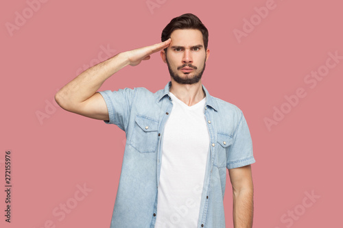Yes sir. Portrait of serious handsome bearded young man in blue casual style shirt standing with salute and looking at camera with attention. indoor studio shot, isolated on pink background.