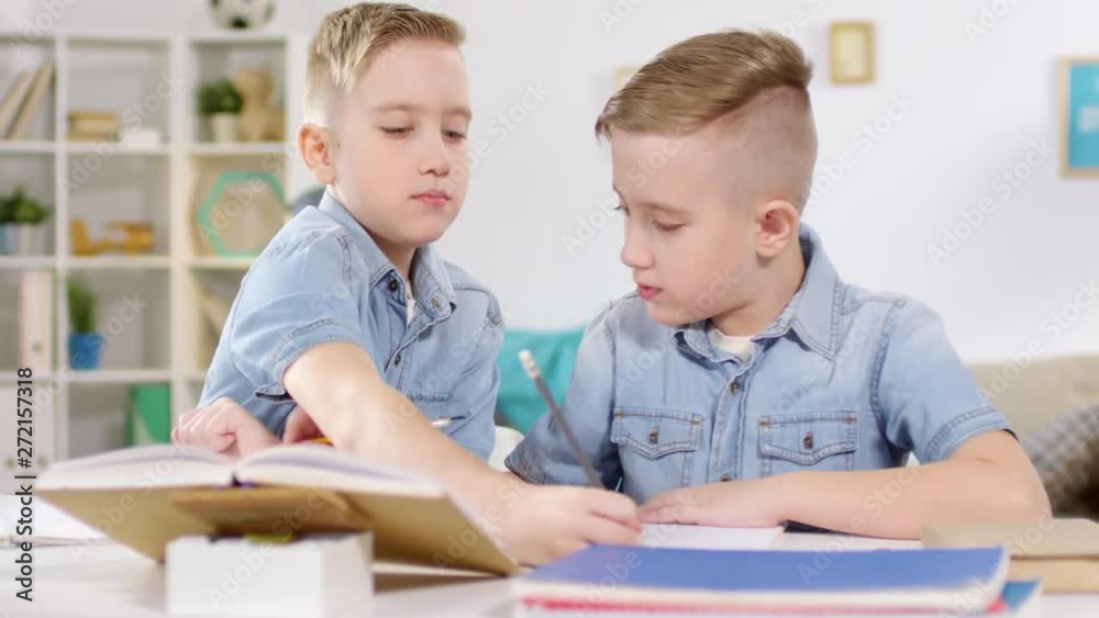Chest-up shot of 10-year-old Caucasian identical twins sitting together at table at home, doing homework and fooling around, writing in each other’s exercise books, then again in their own Chest-up shot of 10-year-old Caucasian identical twins sitting together at table at home, doing homework and fooling around, writing in each other’s exercise books, then again in their own