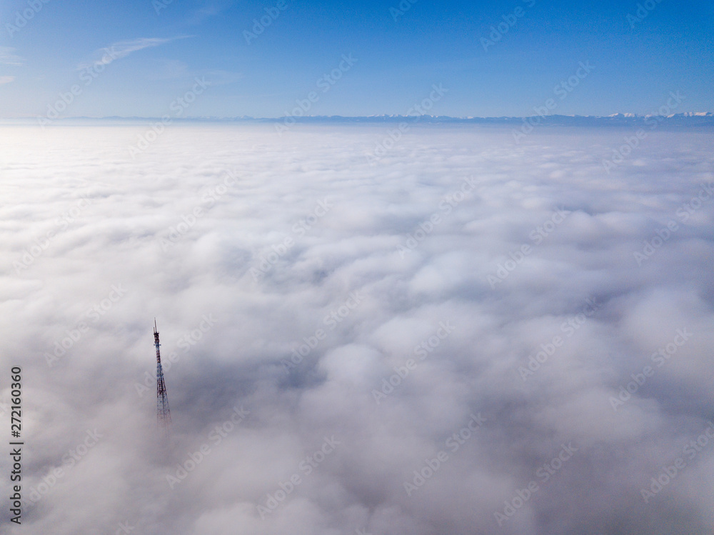Aerial top view of foggy cloudy landscape with radio or TV antenna top on blue sky copy space background and mountain ridge on horizon.