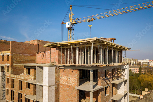 Apartment or office tall building under construction, top view. Brick walls, scaffolding and concrete support pillars. Tower crane on bright blue sky copy space background. Drone aerial photography.