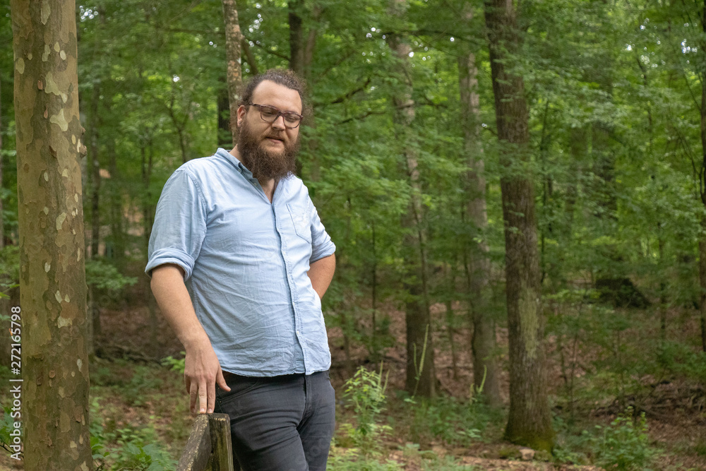 Young bearded man in forest leans against a bridge railing in a thoughtful pose. Relaxed hipster in a natural environment. Young bearded man in forest leans against a bridge railing in a thoughtful pose. Relaxed hipster in a natural environment.