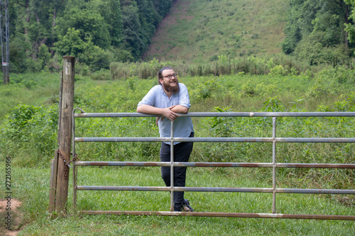 Young bearded man leans against a metal farm gate looking up and smiling. Amused hipster with beard and ponytail in a natural environment. © Jen Wolf