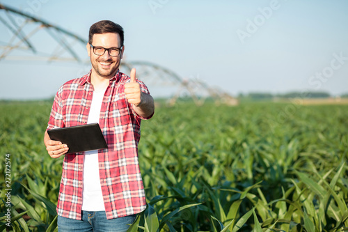 Happy young farmer or agronomist in red checkered shirt showing thumbs up and smiling directly at camera. Standing in corn field, holding a tablet. Organic farming and healthy food production photo