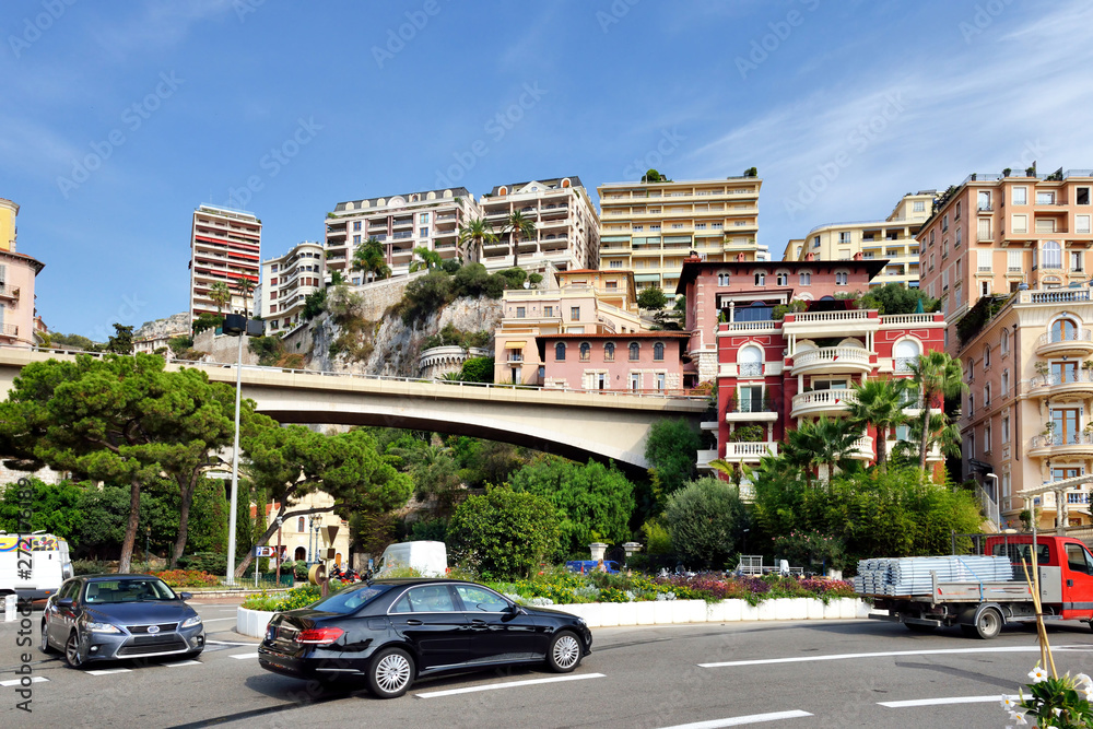 Daylight sunny view to buildings and streets of Monte-Carlo, Monaco. Parked cars and people walking near sidewalk. Bright blue sky with a few clouds on background.