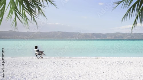 Salda lake like Maldives with white sand and turquoise colored water. One woman sitting on a portable chair near the waters edge, we see some palm leaves at the corner of the frame. Burdur / Turkey. photo