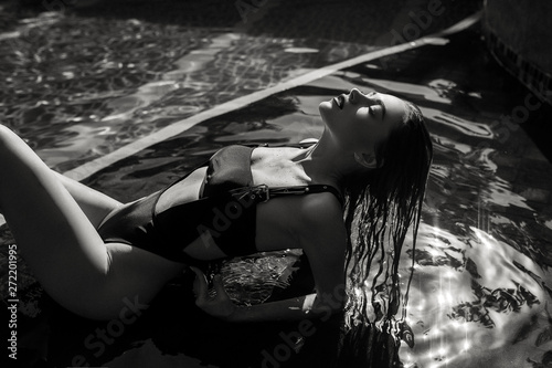 A sexy young girl in a seductive swimsuit is depicted in a black and white photo.  Fashion  Lady with Sports figure  lying on the background of an open summer pool