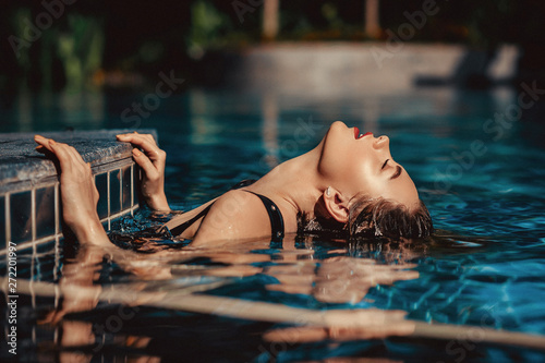 fashion outdoor photo of beautiful woman with blond hair wears luxurious black swimsuit, posing in the swimming pool. Luxury Female lies in the crystal clear pool.