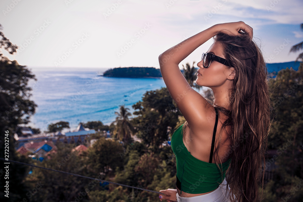 Close up Portrait of beautiful young woman with long hair, wearing elegant white skirt and green top, standing on the edge of the site overlooking the sea beach trees and blue sky. 