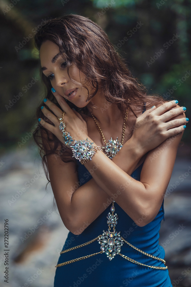Naklejka premium beautiful long- haired brunette girl in bright blue dress and a lot of jewelry on the background of a dark forest. Bracelets, necklaces, silver