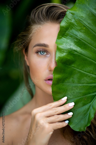 Close up: young woman with natural makeup holds a big green leaf on a blurred green background. Spa and wellness. Youth, teens and skin care concept. Close up