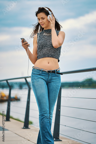 Beautiful young woman with white headphones dances while walking by the river © superelaks