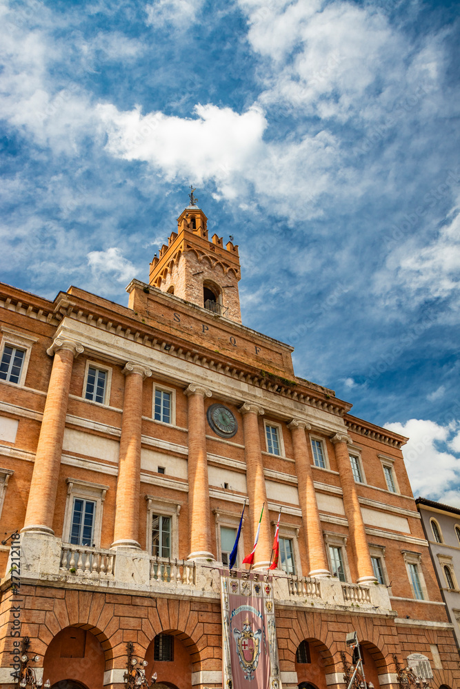 The Town Hall of Foligno in Piazza della Repubblica. The beautiful facade with the colonnade, the clock, the medieval tower and the balcony with the flags of Italy and Europe. Perugia, Umbria, Italy.