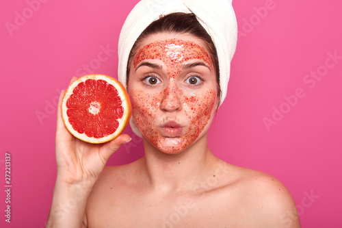 Surprised well kept young woman applied facial colorful scrub, looking directly at camera, holding half of grapefruit in one hand, putting her lips together, stnading half naked in photo studio. photo