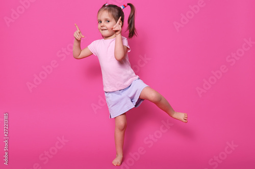 Close up portrait of toddler child baby girl standing in pink casual t shirt and purple short, pointing both fore fingers up and posing on ne leg isolasted over pink background. Childhood concept. photo