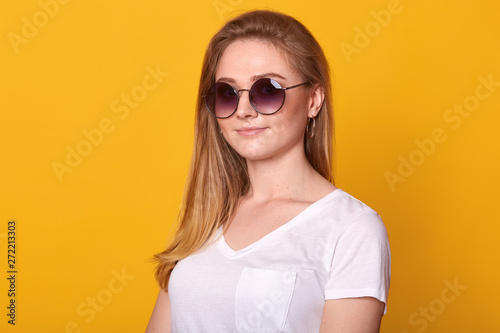 Image of sweet confident young lady standing isolated over bright yellow background in studio, wearing white casual t shirt and fashionable round eyeglasses, having long blond hair. Youth concept.