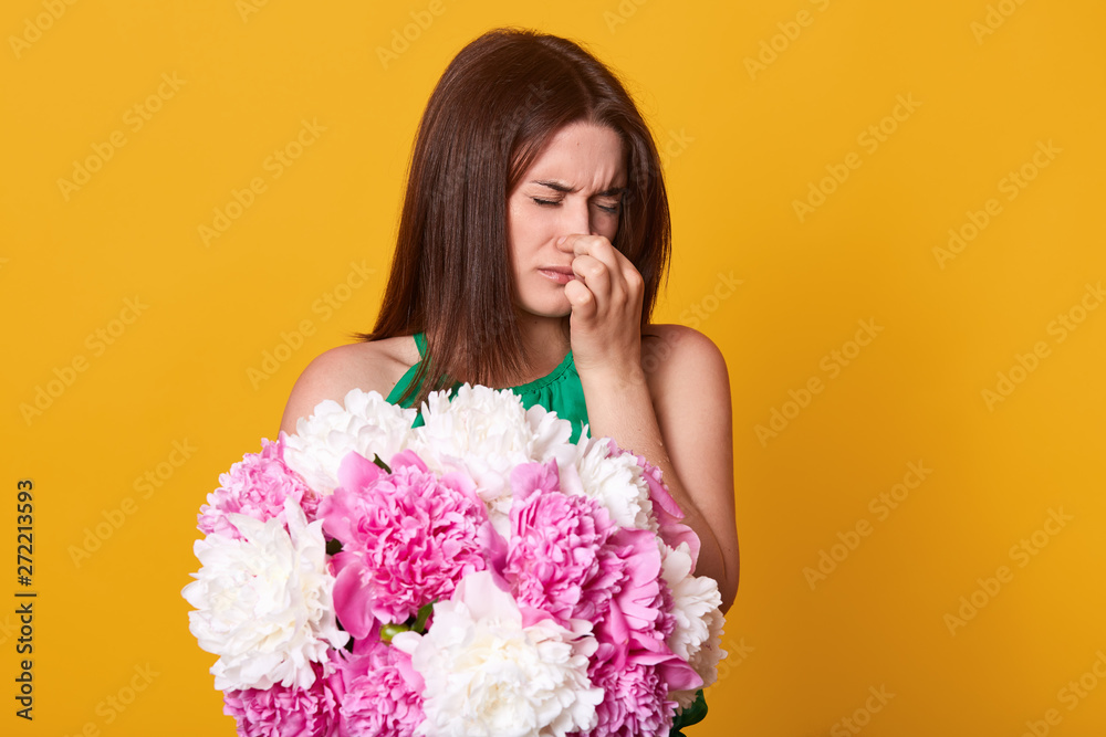 Attractive slender cute model holding her nose, closing eyes, can`t stand smell of peonies, being allergic to flowers, feeling unwell, standing isolated over bright yellow background in studio. Attractive slender cute model holding her nose, closing eyes, can`t stand smell of peonies, being allergic to flowers, feeling unwell, standing isolated over bright yellow background in studio.
