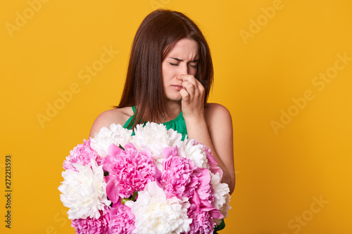 Attractive slender cute model holding her nose, closing eyes, can`t stand smell of peonies, being allergic to flowers, feeling unwell, standing isolated over bright yellow background in studio. photo