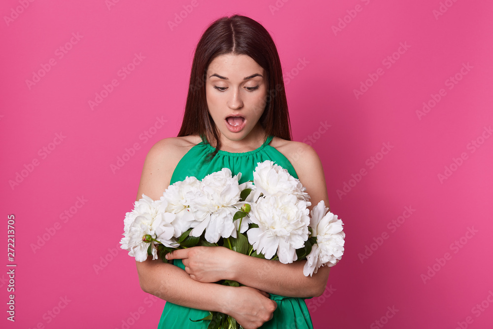 Indoor shot of shocked impressed young woman looking at bouquet with open mouth, stands embracng aring green dress. Unknown hand holding bunch of pink and white peonies.