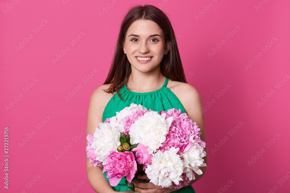 Studio shot of cute brunette European female posing isolated over pink background, looking directly smiling at camera, embracing white and rose peonies with both hands, wearing green sundress.