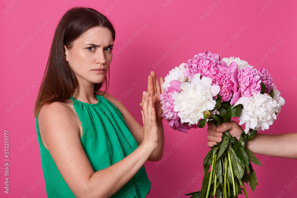 Half length of lady dressed green sundress, refuses beautiful bouquet of white and pink peony flowers from faceless person on yellow studio background, brunette girl with displeased facial expression. Half length of lady dressed green sundress, refuses beautiful bouquet of white and pink peony flowers from faceless person on yellow studio background, brunette girl with displeased facial expression.