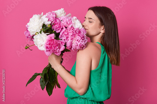 Profile shot of beautiful brunette girl holding with white and rose peonies, attractive woman with big bouquet, female smells flowers, dressed green sundress, posing against yellow pink background. photo