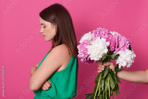 Beautiful girl stands backwards to unknown faceless person who give her peonies isolated over yellow studio background. Young offended woman refuses from flowers from her husband after quarrel. photo