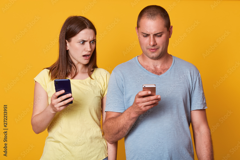 Image of charismatic cute model impressed by her boyfriend messages, looking at screen of his phone, opening mouth and eyes widely with shock, couple holding their smartphones. Relationship concept. Image of charismatic cute model impressed by her boyfriend messages, looking at screen of his phone, opening mouth and eyes widely with shock, couple holding their smartphones. Relationship concept.