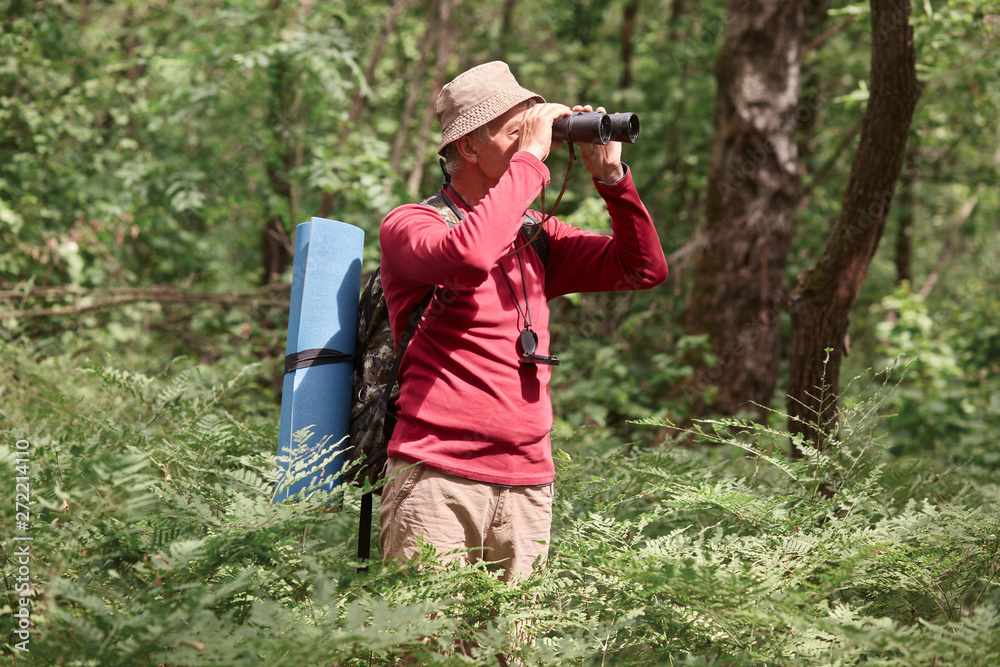 Profile of active hicker looking through binoculars, having compass on his neck, wearing sweatshirt and trousers, taking backpacker and and sleeping pad with him, going camping. Activities concept. Profile of active hicker looking through binoculars, having compass on his neck, wearing sweatshirt and trousers, taking backpacker and and sleeping pad with him, going camping. Activities concept.