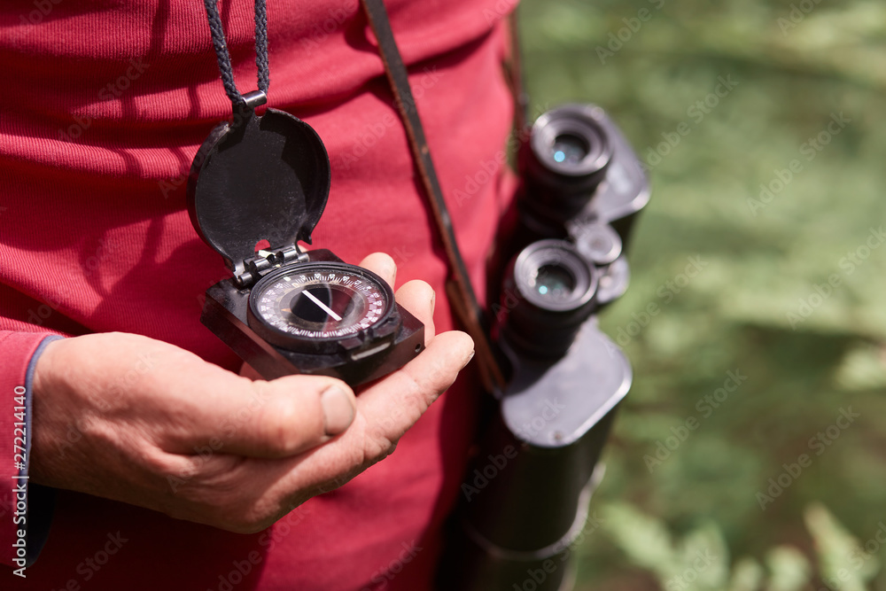 Faceless photo of man's hand with compass, traveler on wooden background wearing red casual shirt, posing with binoculars, finding right way. Traveling, backpacking and active recreaton concept.