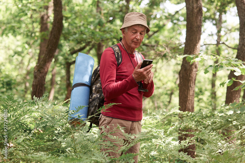 Experienced traveller holding his smartphone in one hand, using device for orientating, being alone, having compass and sleeping pad, wearing hat, trousers, sweatshirt, backpack. Travel concept.