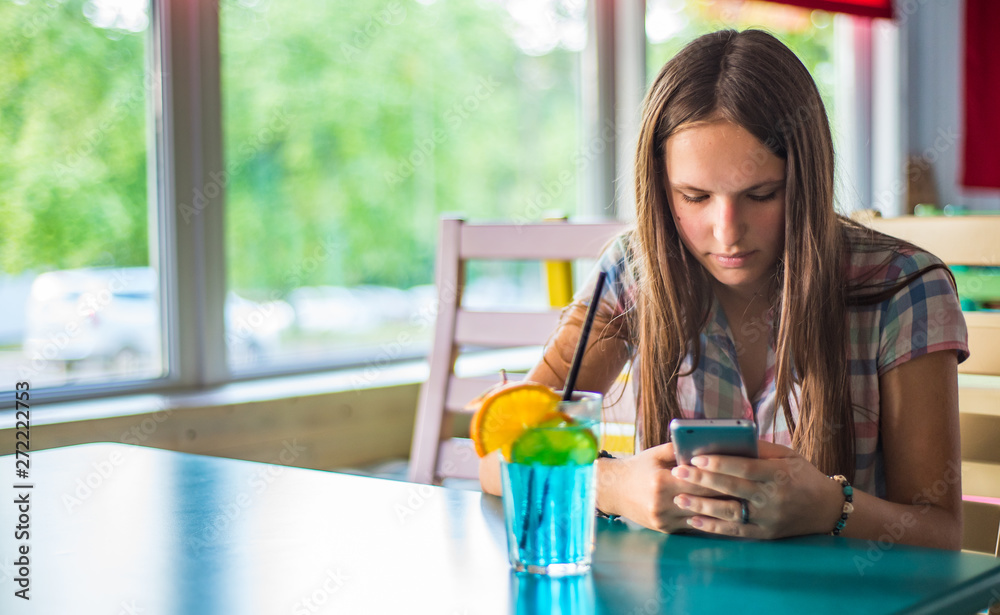 portrait of young teenager brunette girl with long hair sitting indoor in urban cafe, drink a blue lemonade cocktail and use her smartphone portrait of young teenager brunette girl with long hair sitting indoor in urban cafe, drink a blue lemonade cocktail and use her smartphone