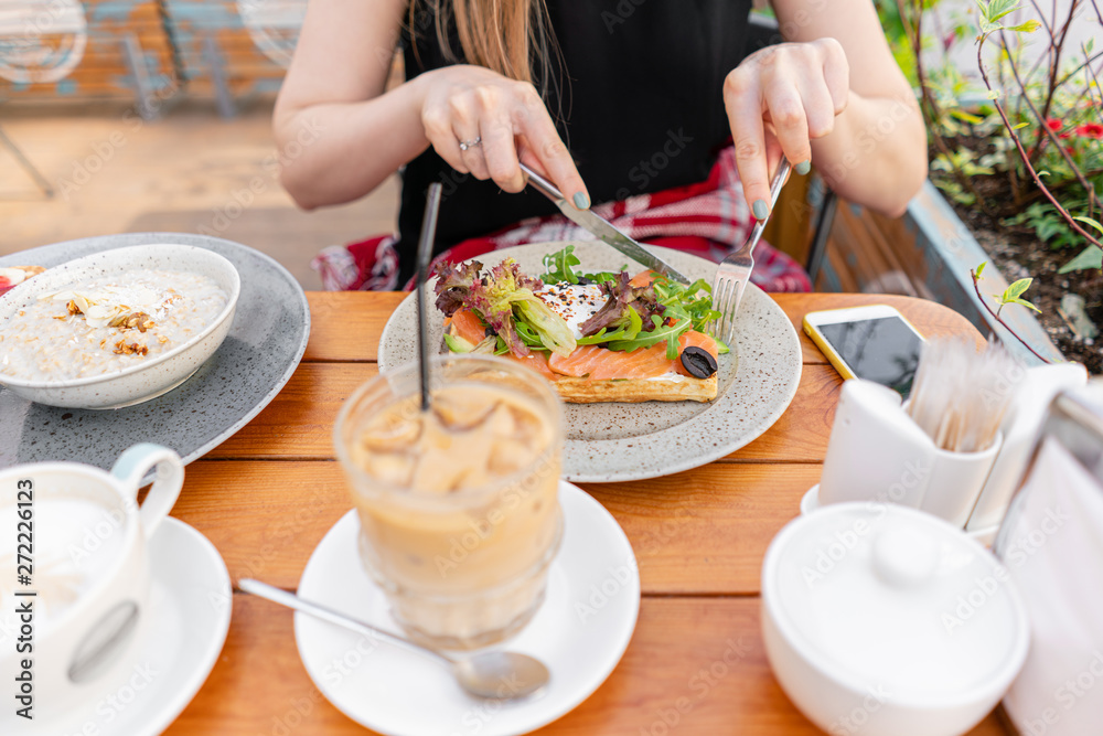 Traditional Belgian waffle with salmon, lettuce leaves and poached egg. A young woman is having Breakfast in a summer cafe, hands with a fork and a knife close-up. Cold coffee with ice cubes