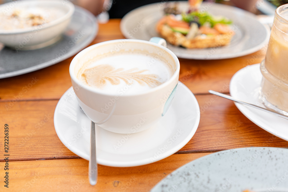 Focus in foreground on coffee cappuccino. A young woman is having Breakfast in a summer cafe. Focus in foreground on coffee cappuccino. A young woman is having Breakfast in a summer cafe.
