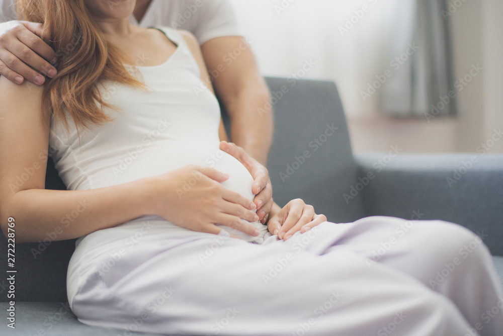 Closed up, Young pregnant Asian woman is sitting on sofa with her husband touch her belly near window which felling happy at family's home. To be new Mother and Father, pregnant woman concept.