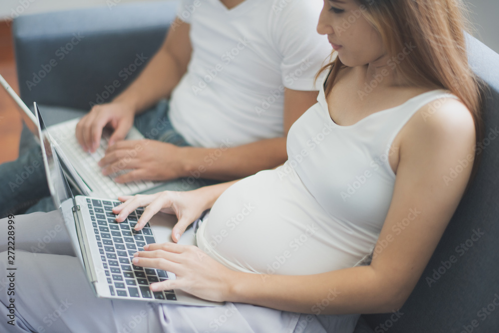 Fototapeta premium Young pregnant Asian woman and her husband are sitting on sofa and working with computer laptop at family's home. To be new Mother and Father, pregnant woman concept.
