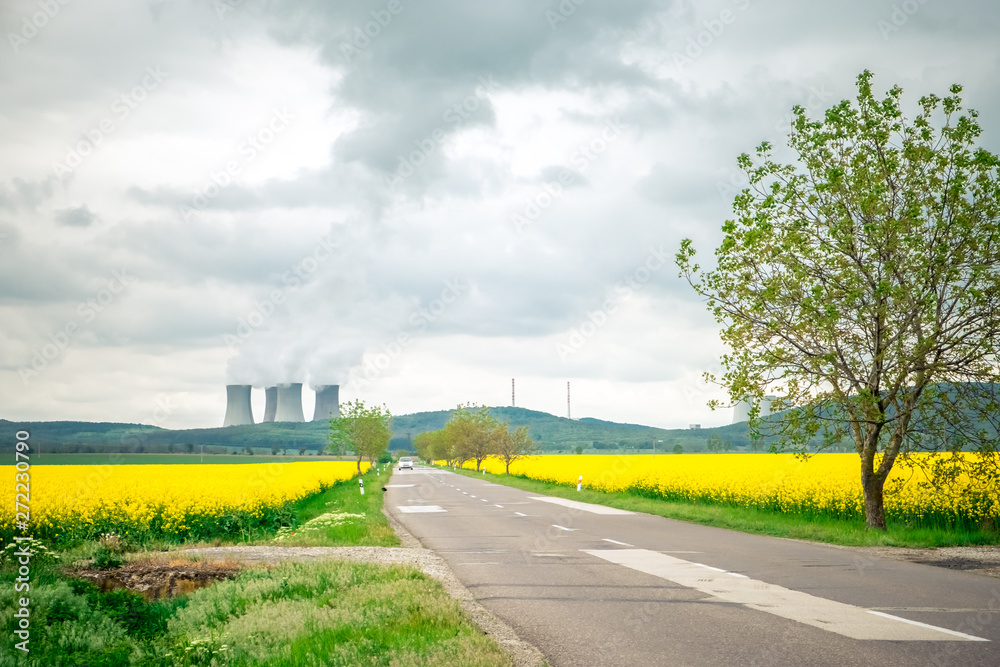 Obraz premium The road between the mustard fields on the background of the pipes of the plant and the cloudy sky
