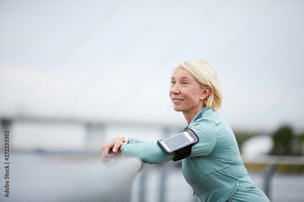 Mature serene sportswoman with blonde hair enjoying solitude while standing in front of riverside after outdoor workout