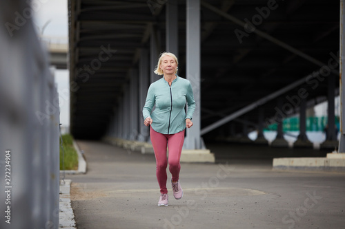 Blonde healthy female in sportswear running down roadway under bridge construction while training in the morning